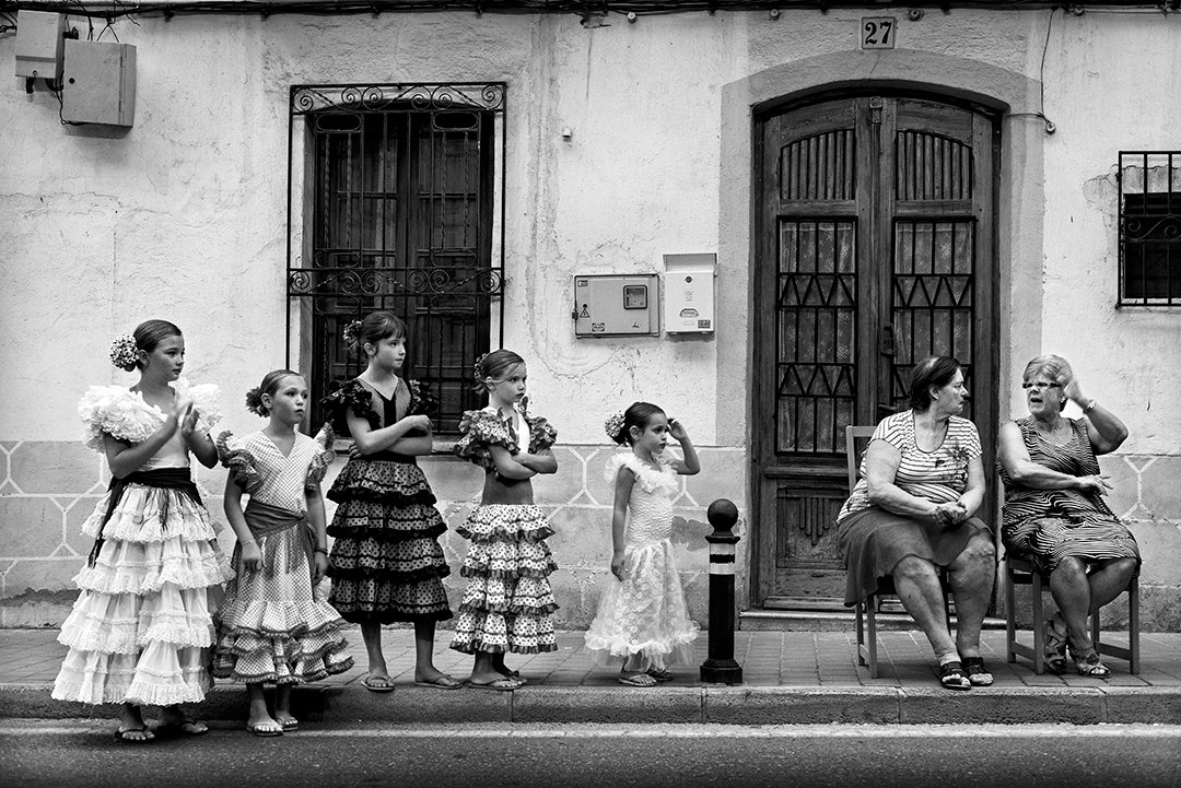 Women in ruffled dresses and seated people in front of a facade.